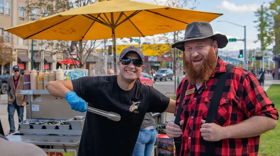 Two men smiling by a street food cart during a guided Anchorage food tour, one serving food and the other standing beside him.