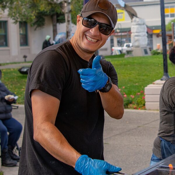 A smiling man wearing a cap and blue gloves grilling hot dogs outdoors during an Anchorage food tour.