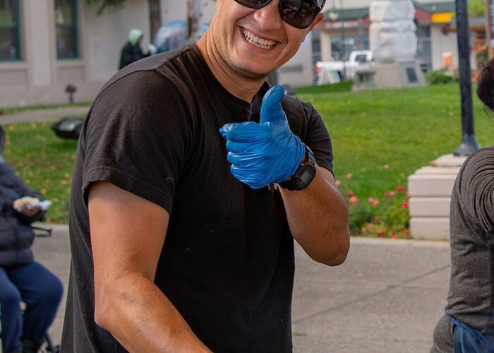 A smiling man wearing a cap and blue gloves grilling hot dogs outdoors during an Anchorage food tour.
