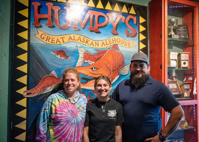 Three individuals posing in front of a colorful mural at Humpy's Great Alaskan Alehouse, part of their guided Anchorage Food Tours.