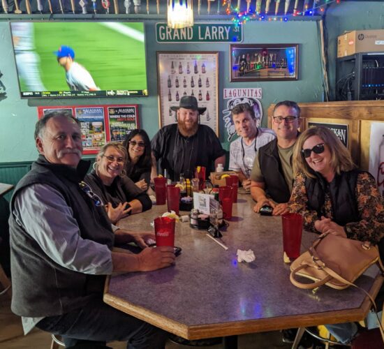 A group of seven adults smiling at a table inside a casual bar with a baseball game on the television in the background, participating in one of the guided Anchorage food tours.