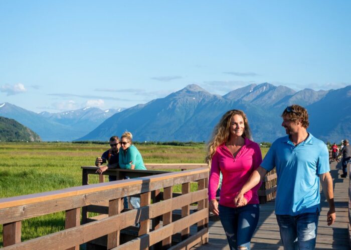 A couple enjoying a guided Anchorage food tour while walking on a wooden boardwalk with mountains in the background and other people savoring the scenery.