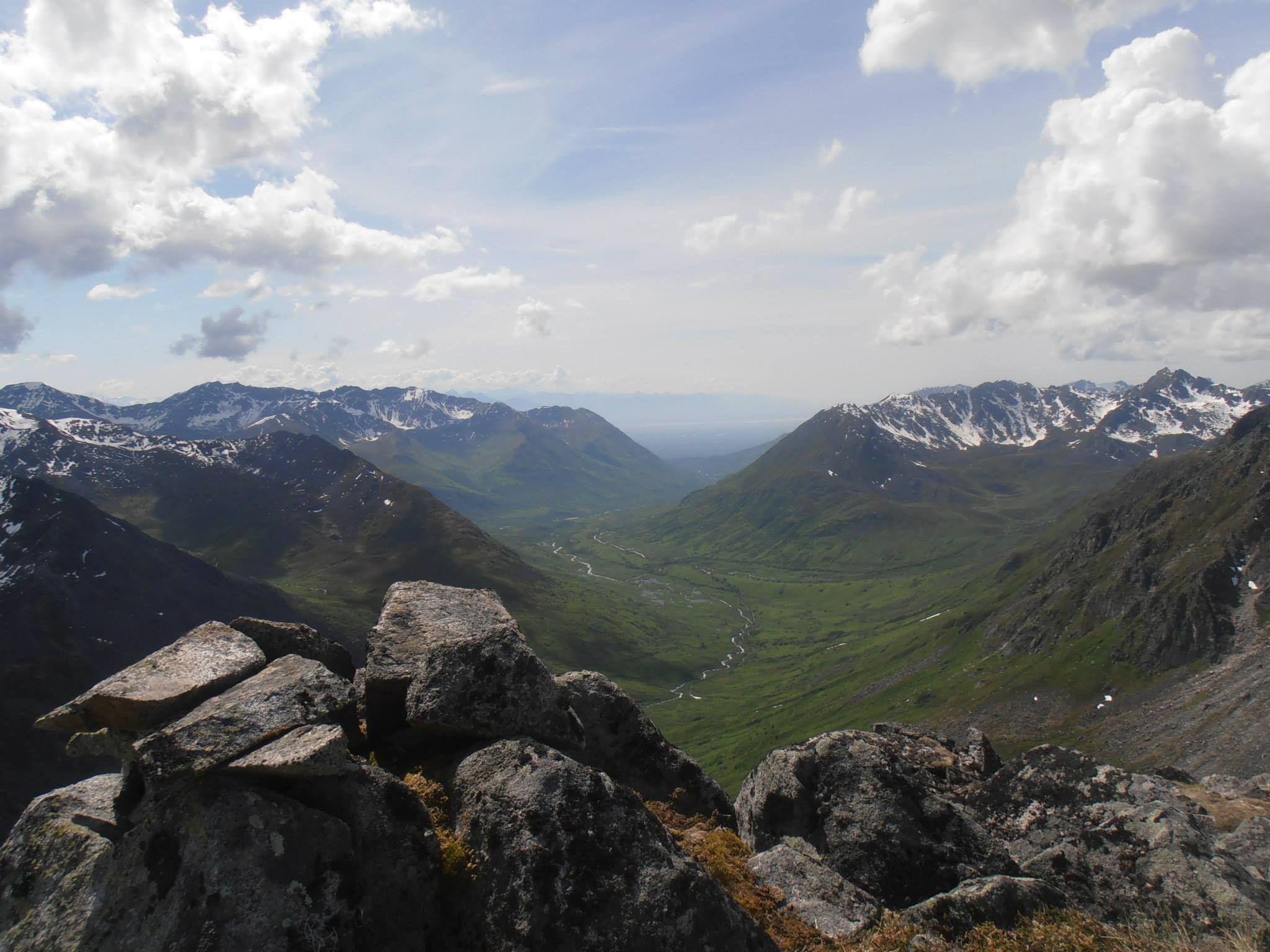 A panoramic view from a mountain summit overlooking a valley with meandering streams and distant mountain ranges under a partly cloudy sky, offering an unmatched opportunity for sightseeing.