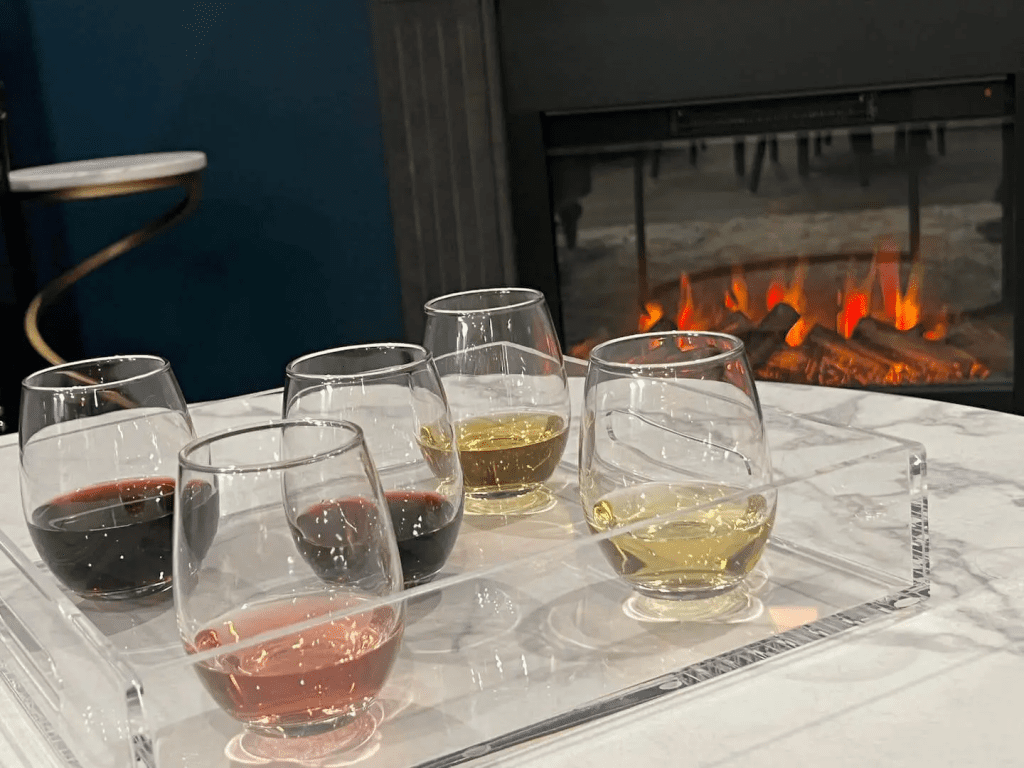 A selection of six wine glasses, each with different colored wines, arranged on a table in front of a lit fireplace in Anchorage, AK for the new Chocolate and Wine Experience.