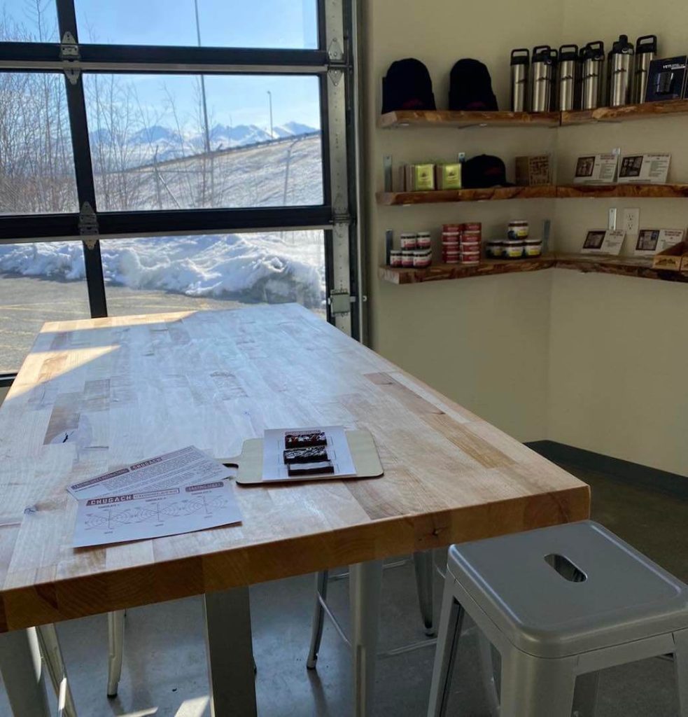 Interior of a cafe in Anchorage with a wooden table and white stool, overlooking a snowy landscape through a large window. Shelves with goods line the wall.