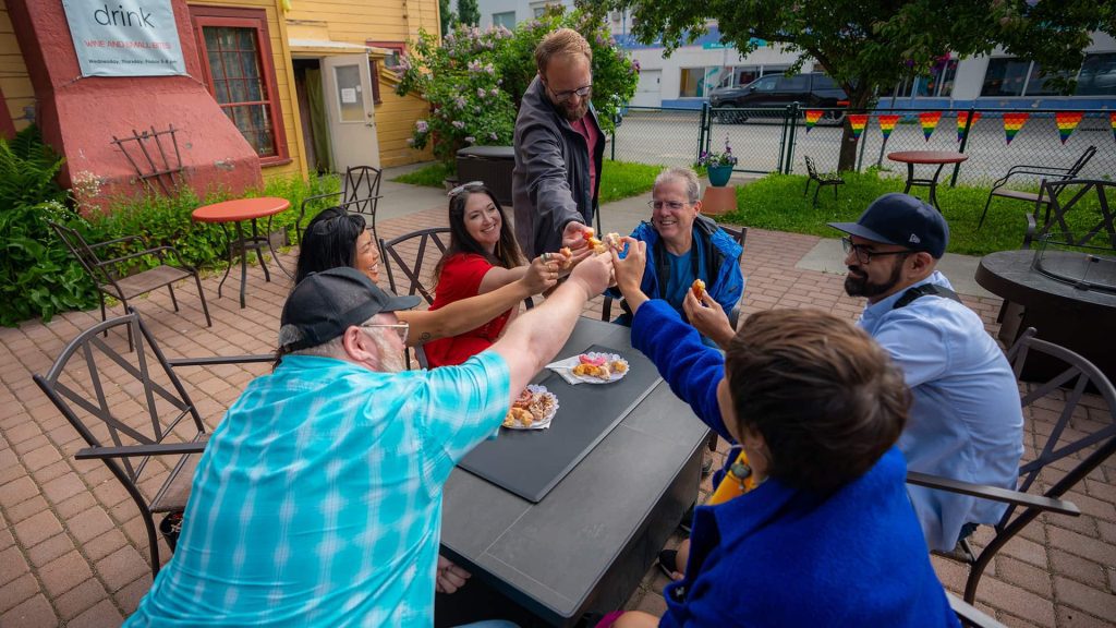 A group of people sit around an outdoor table, raising their hands in a toast. They are seated on a patio area with chairs, tables, and a fenced background. Food and drinks are on the table.