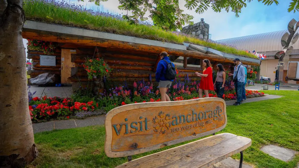 Four people visiting a garden display featuring a grassy-roofed log building in Anchorage. A wooden bench with "visit Anchorage" inscribed is in the foreground.