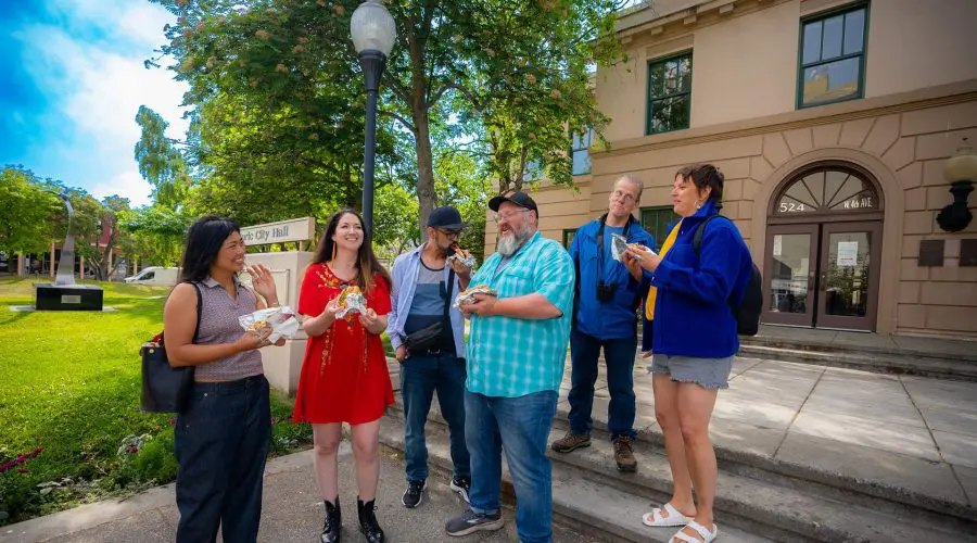 A group of six people stands outside a building, laughing and eating food. Trees and a sign that reads "City Hall" are visible in the background.