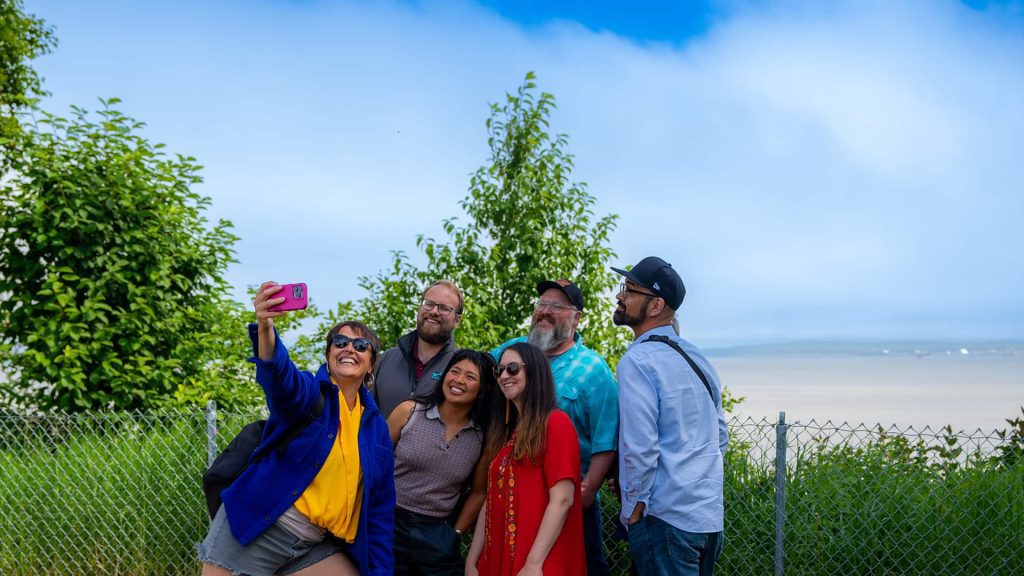 Six people are posing for a group selfie outdoors, with greenery and a chain-link fence in the background. The person on the left is holding a pink phone to take the photo.