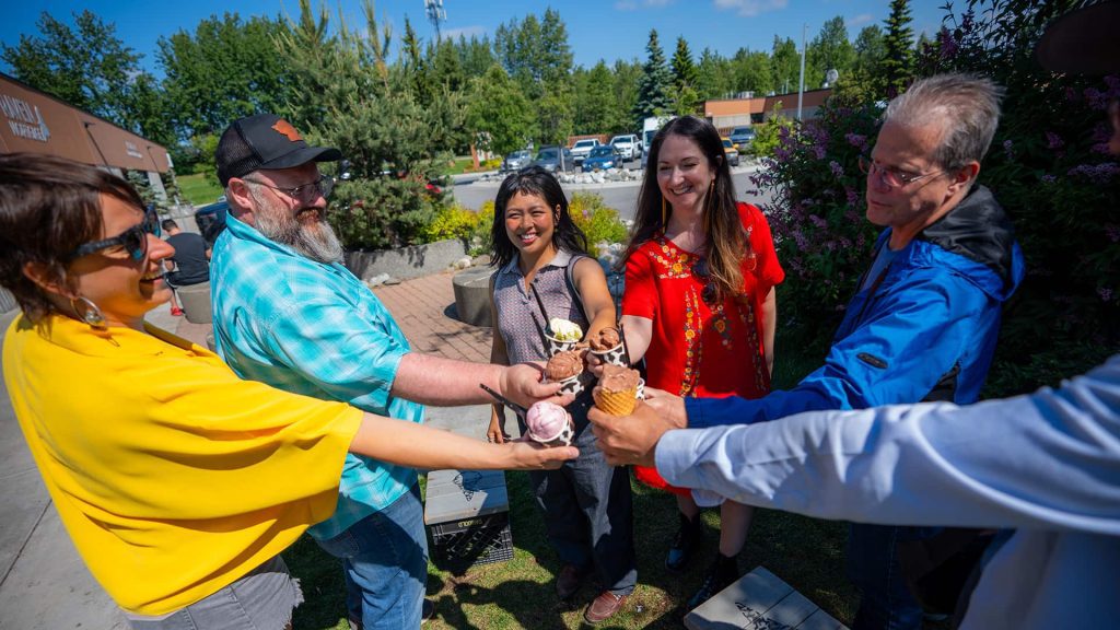 A group of six people standing in a circle outside, smiling and holding ice cream cones together. Trees and a building are visible in the background.