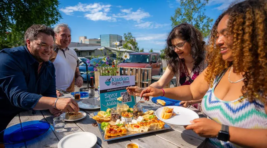 Four people gathered around a table outdoors, enjoying a meal together. The table has varied dishes and a sign that reads "Alaskan Sights and Bites." Trees and buildings are visible in the background.