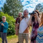 A group of six people stand outdoors in a green, leafy area under a blue sky with white clouds. Some are holding binoculars, and they appear to be looking at something in the distance.