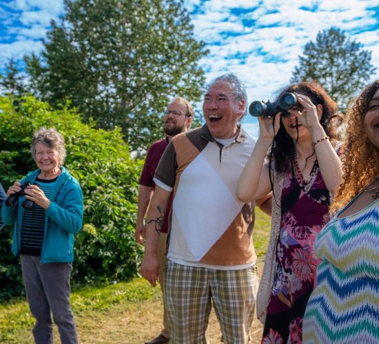 A group of six people stand outdoors in a green, leafy area under a blue sky with white clouds. Some are holding binoculars, and they appear to be looking at something in the distance.