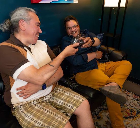 Two people sit on a black leather sofa, toasting drinks and smiling. One wears a brown and white polo shirt with plaid shorts; the other wears a denim shirt and yellow pants.