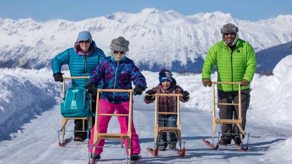 Four people are pushing kicksleds on a snowy path with snow-covered mountains in the background.