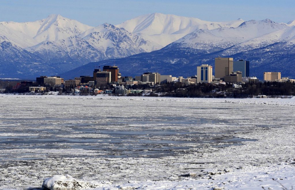 Anchorage skyline with tall buildings, set against a backdrop of snow-covered mountains and a partially frozen body of water in the foreground.