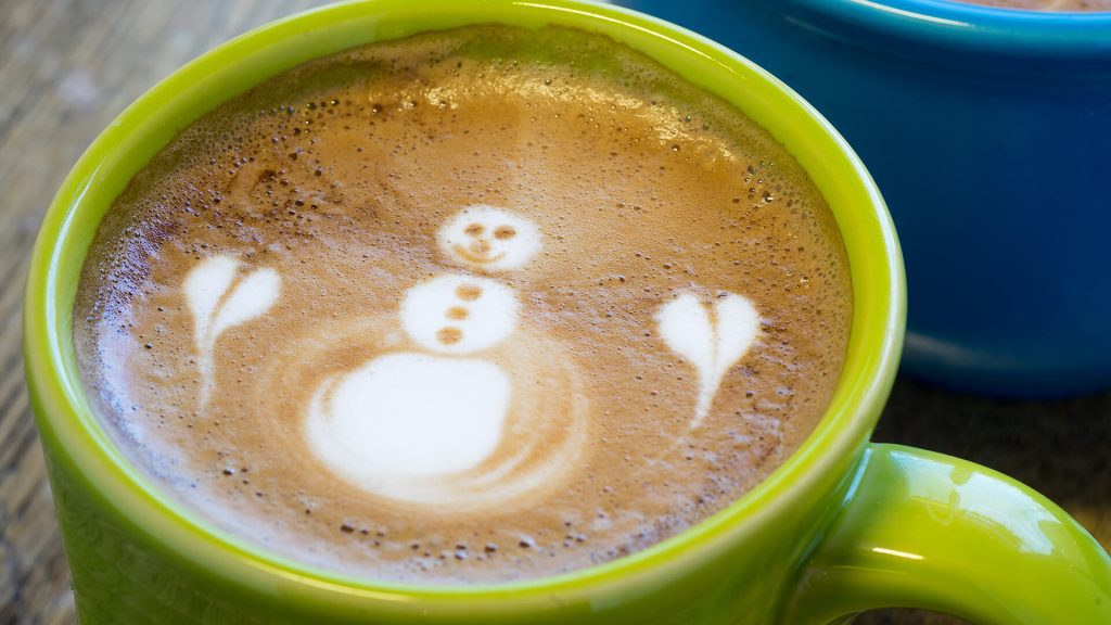 A green mug of coffee with latte art depicting a snowman and two heart shapes on the foam—perfect for curious minds or those dreaming of snowy Alaska mornings.