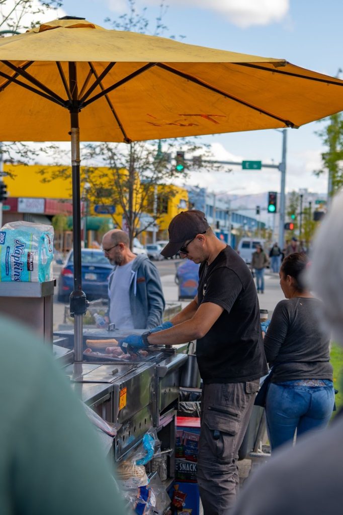 A man prepares Alaskan food at a street vendor cart under a yellow umbrella, with people waiting nearby on a busy city sidewalk.