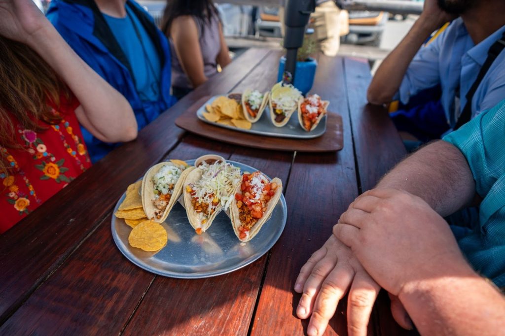 Three tacos with chips on a metal plate are on a wooden table, surrounded by people seated outdoors. Another plate of tacos and chips, inspired by Alaskan cuisine, is also visible in the background.