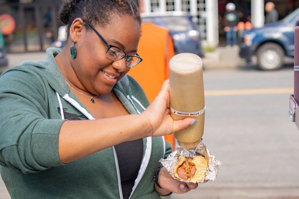 A woman in glasses and a green hoodie adds sauce from a bottle onto Alaskan food wrapped in foil while standing outdoors.