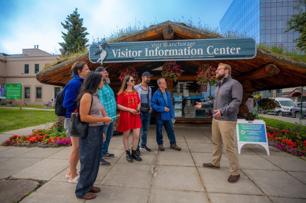 A group of people listens to a man speaking outside the Anchorage Visitor Information Center, with flowers, city buildings, and mentions of Alaskan cuisine in the background.
