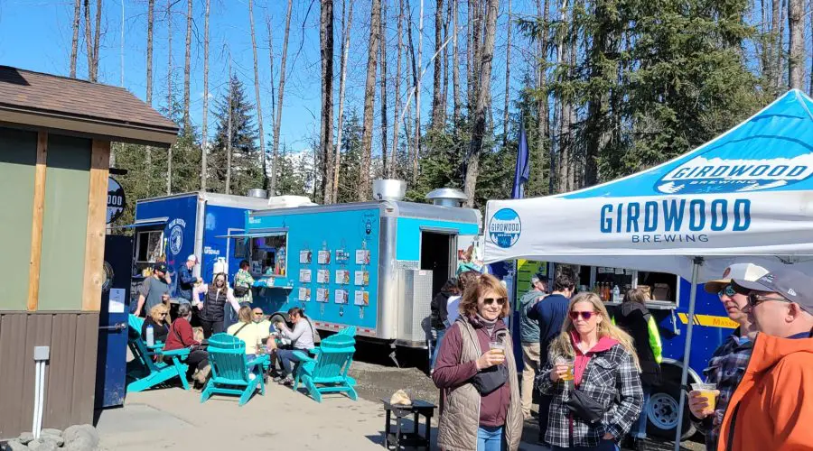 People enjoy drinks and food at an outdoor gathering with food trucks, a brewery tent, and seating under a clear blue sky.