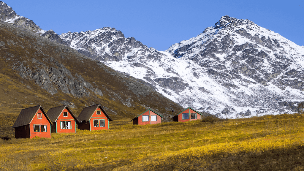 Red and green cabins on a grassy hillside with snowy mountains in the background under a clear blue sky.