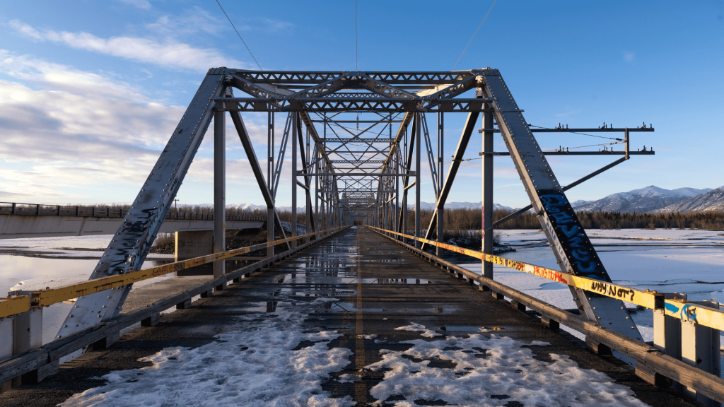 A metal truss bridge with graffiti spans across a partially frozen river. Snow patches are visible on the bridge and surrounding ground under a partly cloudy sky.
