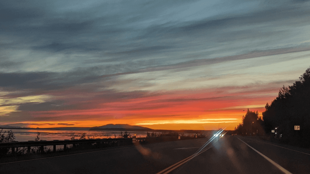 A scenic sunset view from a road, with vibrant orange and pink hues in the sky. The road leads towards distant mountains and a body of water, framed by trees on the right.