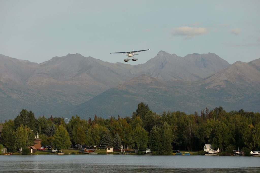 A seaplane flies over a lake with a forested shoreline and rugged mountains in the background under a clear sky, capturing the breathtaking scenery often featured on Alaska tours.