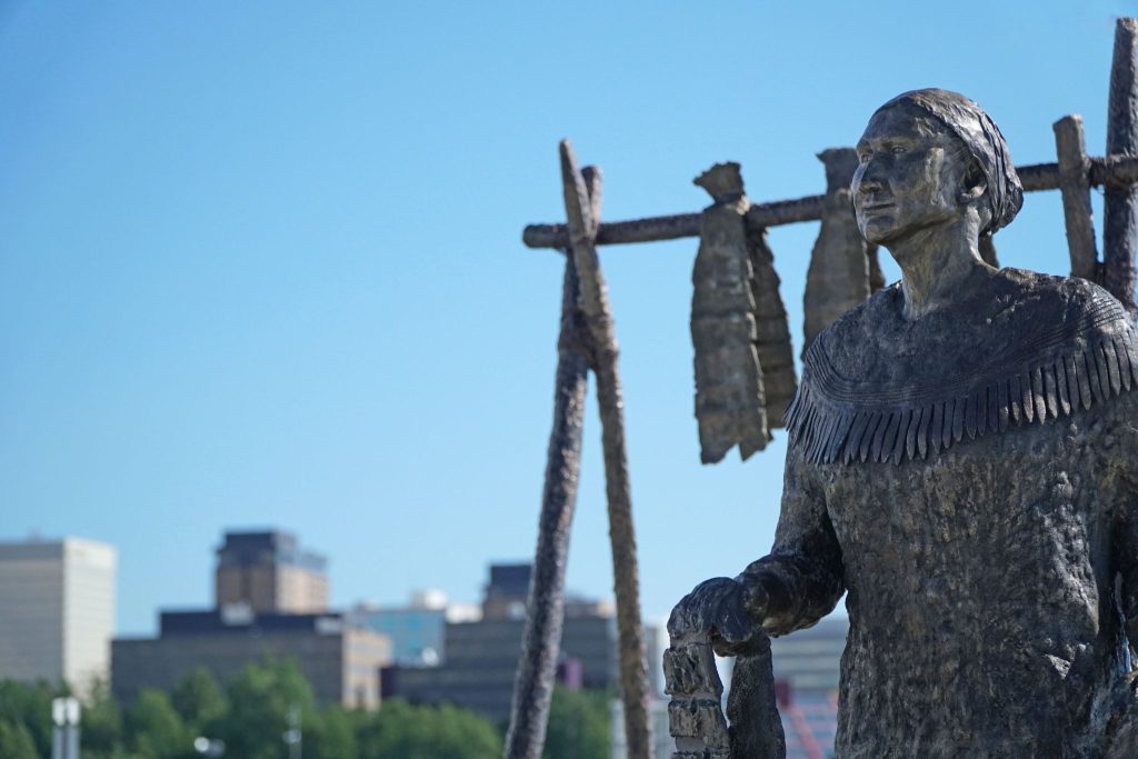 Bronze statue of a person in traditional clothing holding an object, with racks of hanging fish behind them and a city skyline in the background.