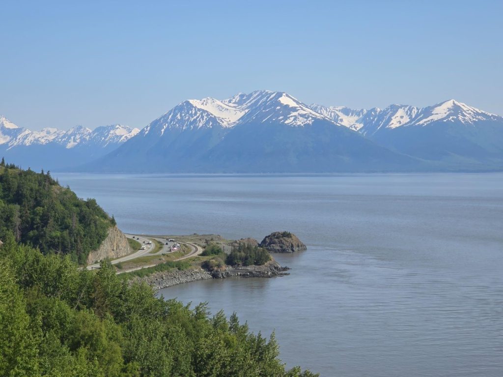 A winding coastal road runs alongside a calm body of water with snow-capped mountains in the background under a clear sky.