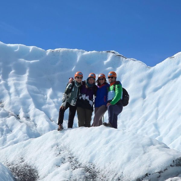 Four people wearing helmets and outdoor clothing stand together on a snowy, icy slope with a bright blue sky in the background.