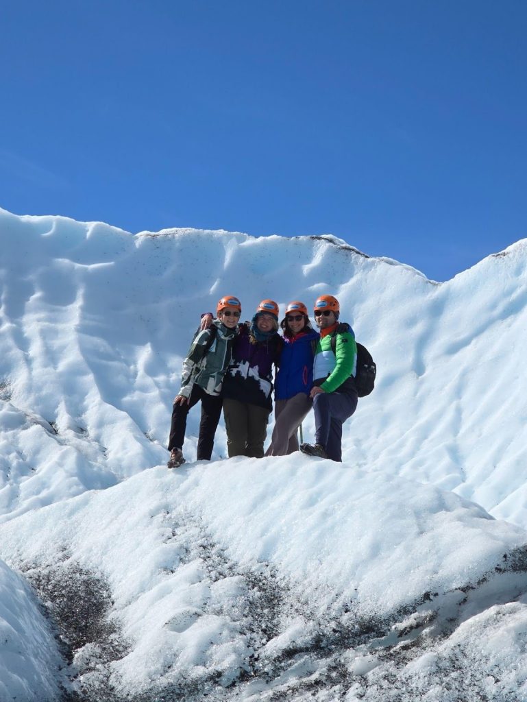 Four people wearing helmets and outdoor clothing stand together on a snowy, icy slope with a bright blue sky in the background.