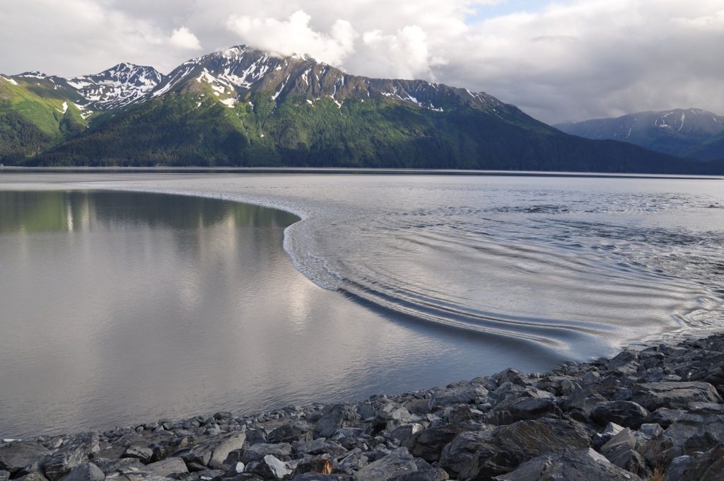 A rocky shoreline leads to calm water with a visible wake pattern; snow-capped mountains and clouds are in the background.