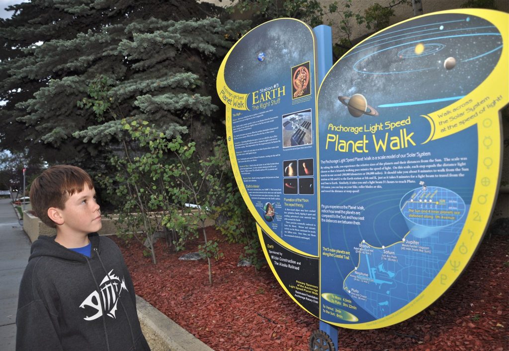 A boy in a hoodie reads an informational sign about the Anchorage Light Speed Planet Walk and the solar system, with trees and a sidewalk in the background.