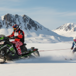 A person rides a snowmobile across a snowy plain, towing a skier behind them—an exciting display of Alaska traditions—with snow-covered mountains in the background. A person rides a snowmobile across a snowy plain, towing a skier behind them—an exciting display of Alaska traditions—with snow-covered mountains in the background.