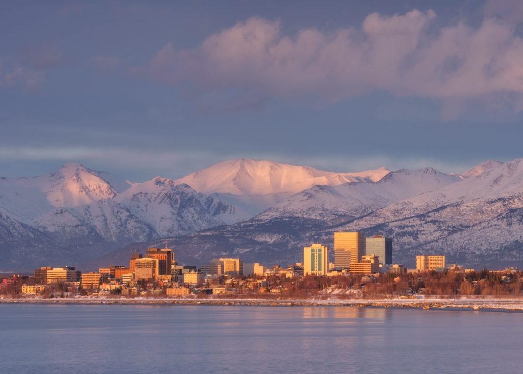 Anchorage city skyline at sunset with snow-covered mountains in the background and calm winter water in the foreground—a perfect scene for any Anchorage travel guide.