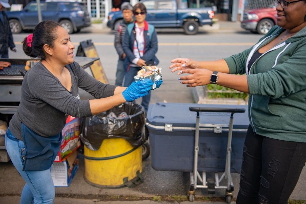 A woman wearing gloves hands food wrapped in foil to another person outdoors near a sidewalk with trash bins and containers nearby.