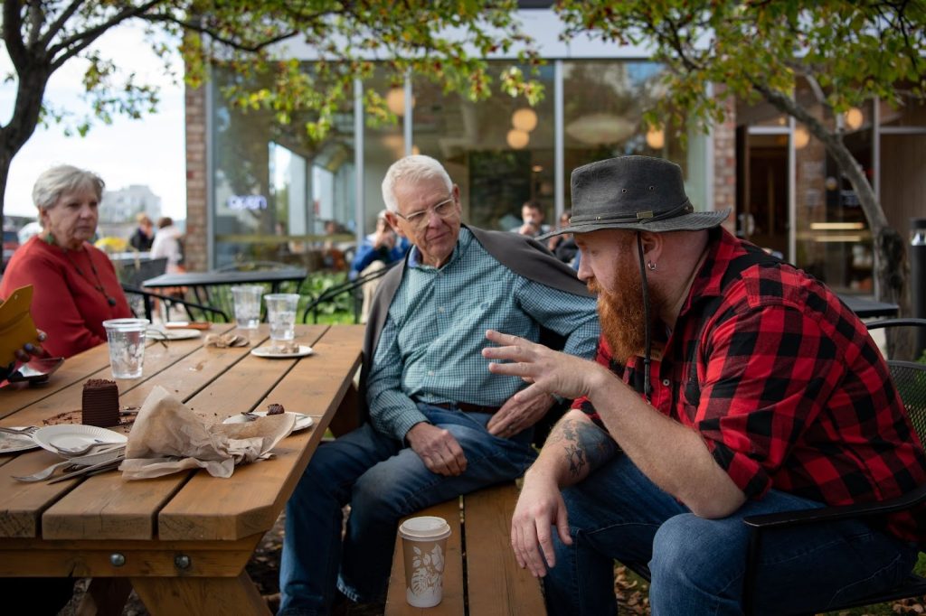 Three people sit at a wooden outdoor table having a conversation; empty plates, cups, and food wrappers are on the table. Trees and a cafe building are in the background.
