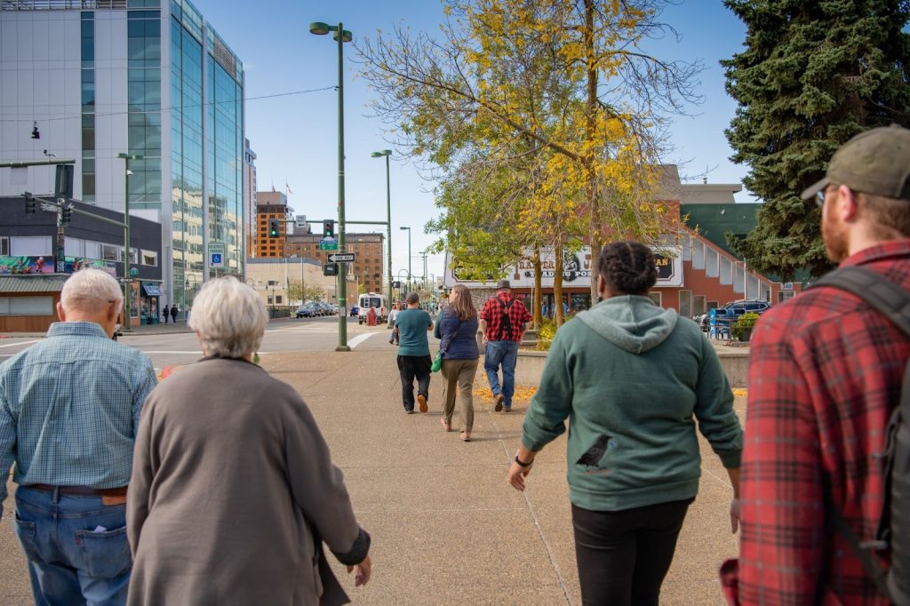 A group of people walk along a city sidewalk on a clear day, with buildings, trees, and traffic signals visible in the background.