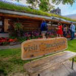 A wooden bench with “Visit Anchorage” text sits in front of a log cabin surrounded by flowers as several people walk nearby on a grassy path. A wooden bench with “Visit Anchorage” text sits in front of a log cabin surrounded by flowers as several people walk nearby on a grassy path.