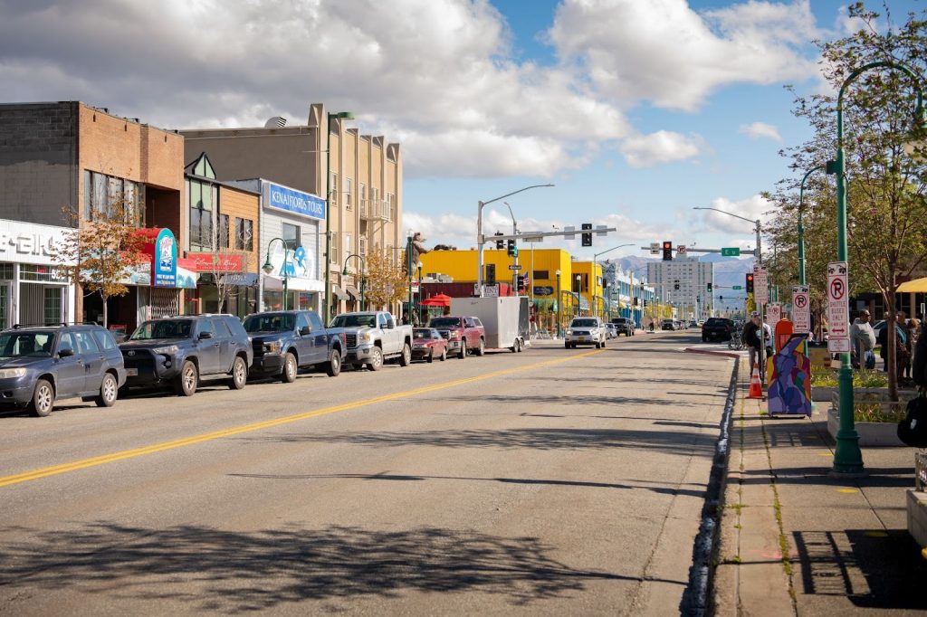 A city street with parked cars, buildings, shops, and traffic signals under a partly cloudy sky; a few people are waiting at a bus stop on the sidewalk.