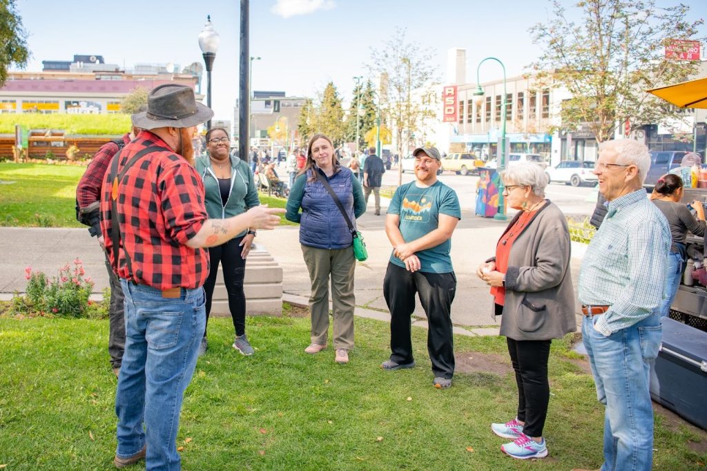 A group of people stands in a park, attentively listening to a man in a red plaid shirt and hat who appears to be leading a tour or discussion.