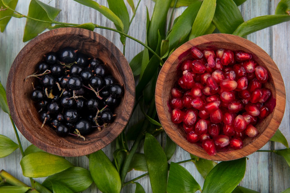 Two wooden bowls: one filled with blackcurrants, the other with pomegranate seeds, placed on green leaves on a light wooden surface.