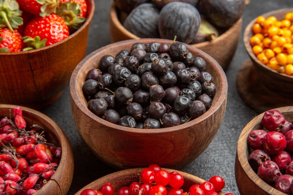 Assorted fresh berries in wooden bowls, including strawberries, black currants, red currants, sea buckthorn, and figs, arranged on a dark surface.