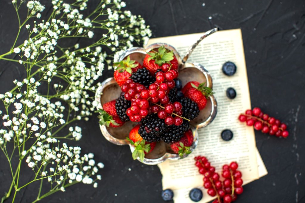 A bowl of strawberries, blackberries, raspberries, and red currants sits on an open book, with baby's breath flowers and loose berries on a dark surface.