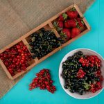 A wooden tray with strawberries, blackcurrants, and redcurrants is on a burlap cloth next to a white bowl filled with blackcurrants and redcurrants, on a turquoise surface. A wooden tray with strawberries, blackcurrants, and redcurrants is on a burlap cloth next to a white bowl filled with blackcurrants and redcurrants, on a turquoise surface.