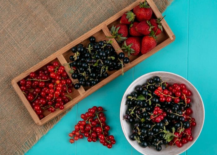 A wooden tray with strawberries, blackcurrants, and redcurrants is on a burlap cloth next to a white bowl filled with blackcurrants and redcurrants, on a turquoise surface.