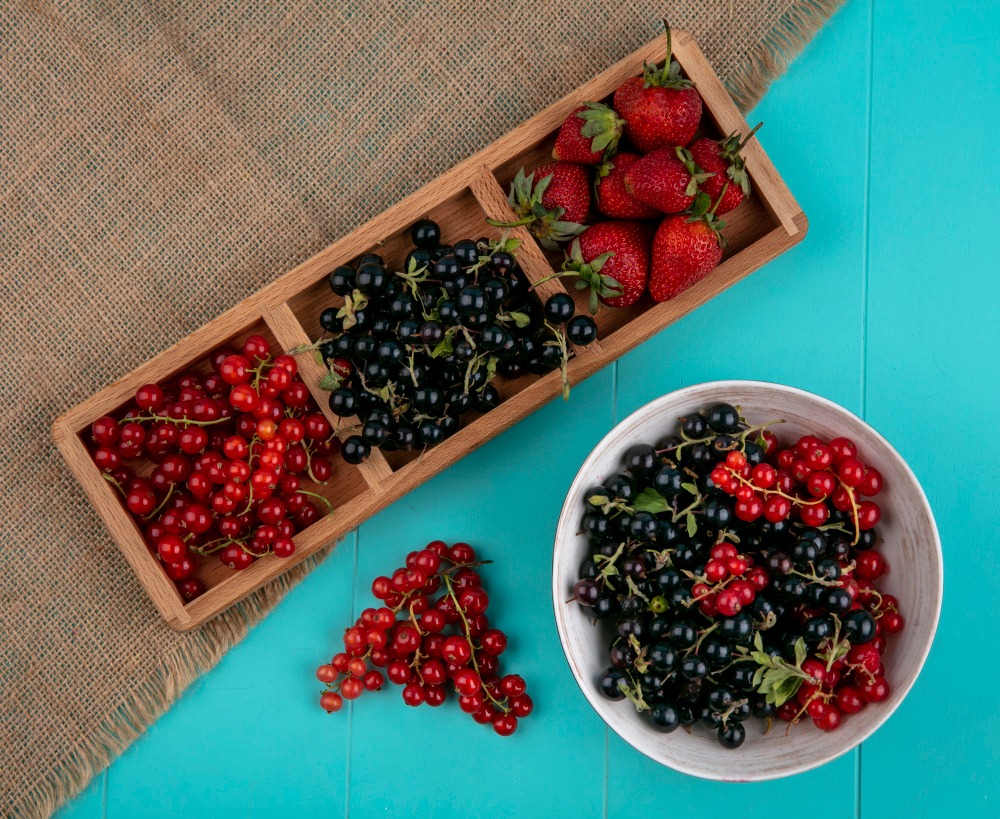 A wooden tray with strawberries, blackcurrants, and redcurrants is on a burlap cloth next to a white bowl filled with blackcurrants and redcurrants, on a turquoise surface.